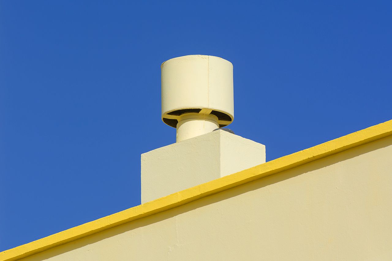 our-story-01 Yellow industrial chimney against a vibrant blue sky, representing modern architecture.